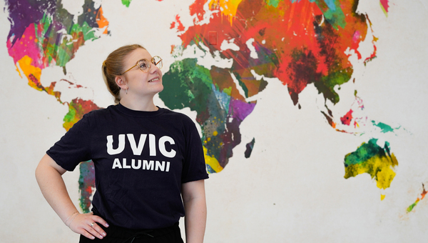 A happy person with a UVic Alumni T-shirt standing in front of a colourful world map