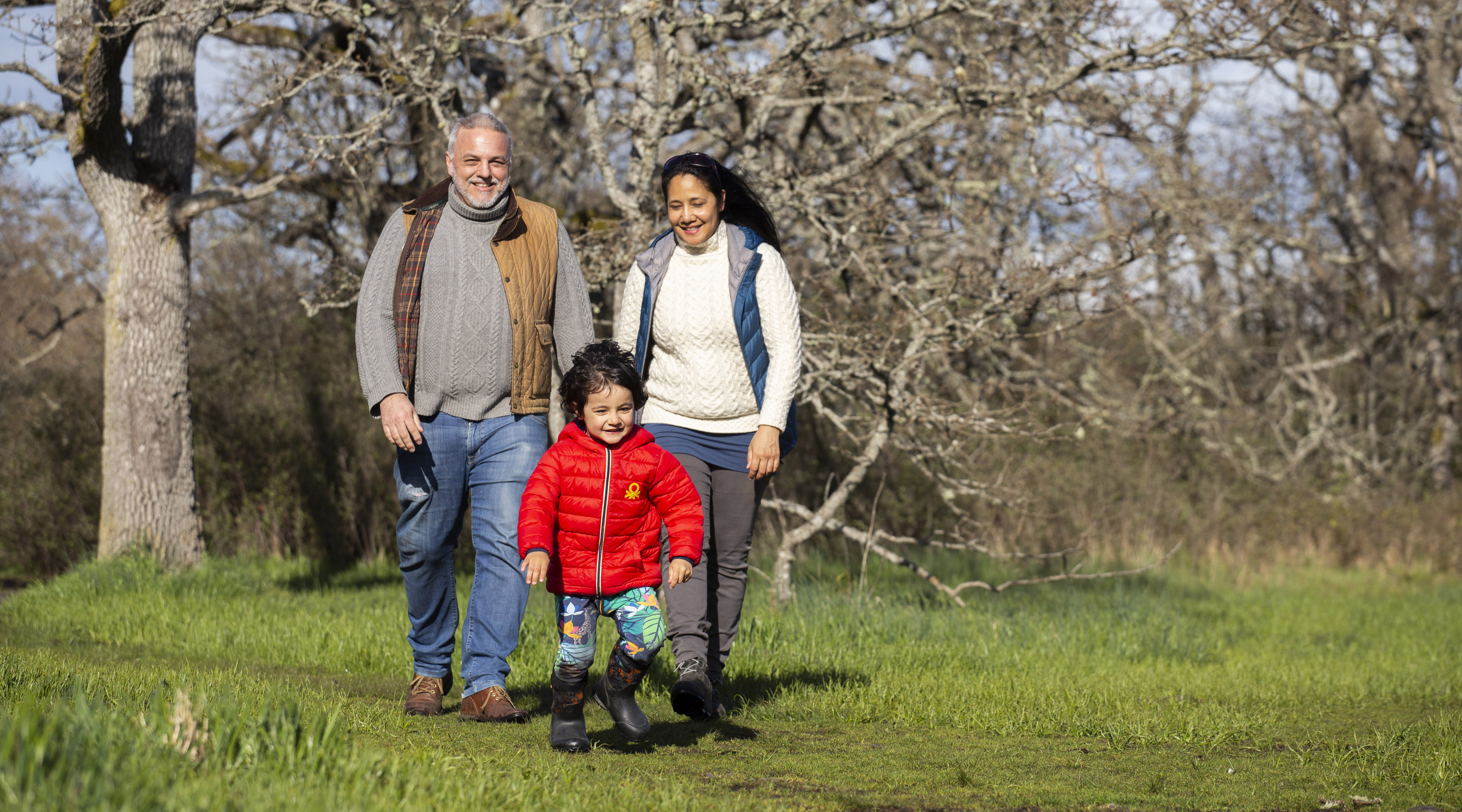 A man, woman, and small child walk in a nature park surrounded by bare trees.